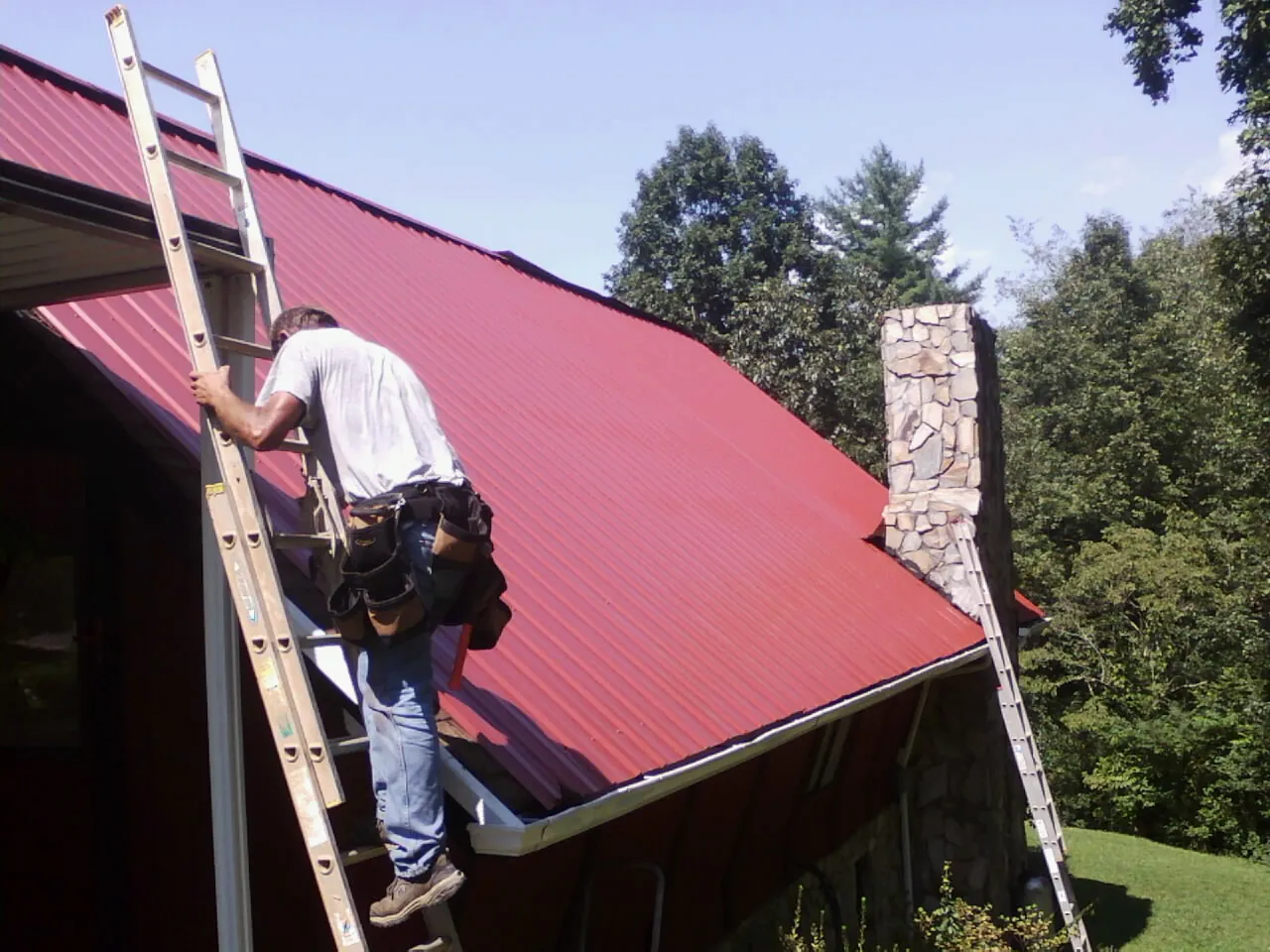 Roofer on a ladder beside a red metal roof and stone chimney — Soffit & Fascia Repair by Roofing Brothers in Seattle